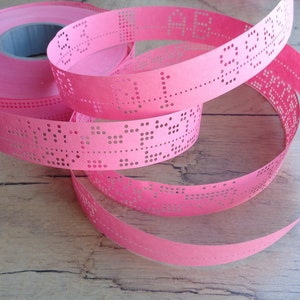 Può includere: Un rotolo di nastro di carta rosa brillante con una serie di fori punzonati disposti in motivi. Il nastro è avvolto, mostrando sezioni di carta con punti dati perforati e un nucleo centrale in plastica grigia visibile nel rotolo.