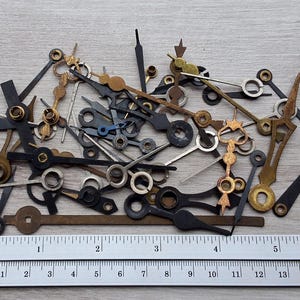Op de afbeelding: Een verzameling vintage uurwerk wijzers in diverse vormen en maten, in zwart, goud en zilver. Een liniaal met inch- en centimeter maten is inbegrepen, waaruit blijkt dat de wijzers tot 15 cm lang zijn. De wijzers liggen op een licht houten oppervlak.