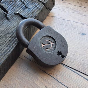 May include: A vintage, weathered metal padlock with a curved handle. The lock is dark gray with a circular design and keyhole. The background features a wooden surface and a dark, textured wooden block.