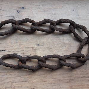 Puede incluir: Una sección de una cadena de hierro oxidado con eslabones ovalados. La cadena es de color marrón oscuro y parece desgastada, con una textura rugosa. Los eslabones están entrelazados, formando una longitud flexible de cadena.