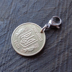 May include: A gold-colored Ukrainian coin with the year 2001 and the word "Ukraina" inscribed on it. The coin features the Ukrainian coat of arms, a trident, and wheat stalks. The coin is attached to a silver-colored metal key chain.