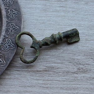 Può includere: Una chiave in metallo verde scuro invecchiato con un arco a forma di cuore e un gambo cilindrico testurizzato, posizionata accanto a una parte di un piatto circolare in metallo argentato con un motivo decorativo in rilievo sul bordo.