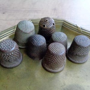 Puede incluir: Una colección de ocho dedal de metal vintage en varios tonos de marrón y gris. Los dedales están dispuestos en una bandeja de latón con un borde decorativo.