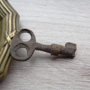 Può includere: Una chiave di metallo arrugginita con un design a doppio foro e un'estremità arrotondata. La chiave è appoggiata su una superficie di legno.