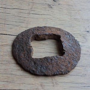 Könnte beinhalten: Eine kreisförmige Unterlegscheibe aus Metall mit einem quadratischen Mittelloch. Das Objekt hat eine stark oxidierte, dunkelbraune und strukturierte Oberfläche mit sichtbaren Anzeichen von Korrosion und Abnutzung am gesamten Rand.