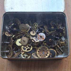 Op de afbeelding: Een open metalen doos vol kleine uurwerktandwielen. De tandwielen variëren in grootte en kleur, met messing en zilver tinten. Sommige tandwielen hebben zichtbare tanden, andere ingewikkelde ontwerpen.