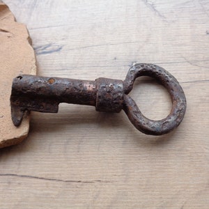 May include: Close-up of a vintage, rusty metal key with a circular handle. The key has a worn, aged appearance, suggesting it is an antique. The key is resting on a light brown wooden surface, with a piece of broken terracotta tile.
