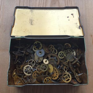 Op de afbeelding: Een open, rechthoekige metalen doos gevuld met een verzameling kleine uurwerktandwielen van messing en zilver. De doos heeft een goudkleurige binnenkant en een groene buitenkant, rustend op een houten oppervlak.