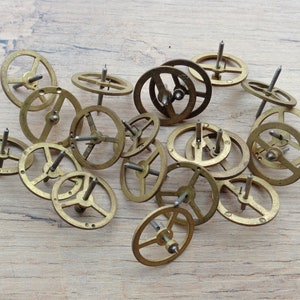 Op de afbeelding: Een verzameling van 15 kleine, messingkleurige uurwerkjes met een centrale pen. De tandwielen zijn willekeurig gerangschikt op een lichtbruin houten oppervlak.