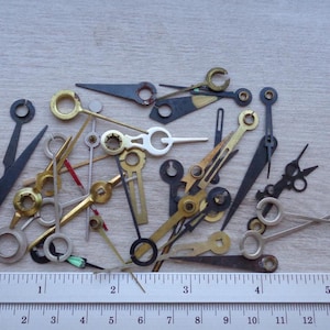 Op de afbeelding: Een verzameling vintage uurwerk wijzers in diverse vormen en kleuren, waaronder goud, zwart en zilver. Een liniaal met inch- en centimeter maten staat onderaan. De langste wijzer is ongeveer 5 cm lang.