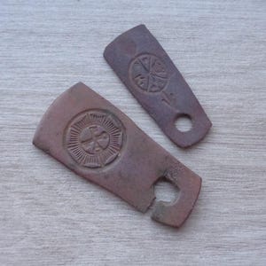 May include: Two antique, reddish-brown, carved wooden pieces with circular and cross designs. One piece has a broken end with a hole, the other has a hole at the end. The pieces are on a light wood surface.