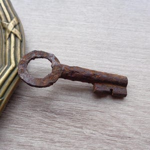 May include: A rusty metal key with a round head and a long, narrow shaft. The key is lying on a wooden surface.