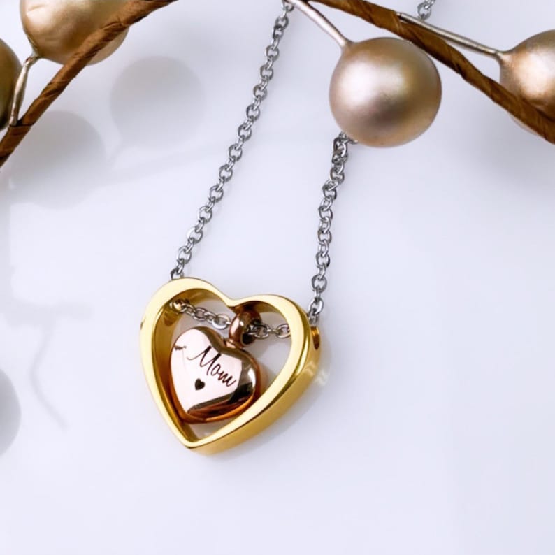 Pode incluir: Um colar com pingente em forma de cora&ccedil;&atilde;o dourado com um cora&ccedil;&atilde;o menor em ouro rosa no interior. O cora&ccedil;&atilde;o menor tem a palavra "Mom" gravada. A corrente do colar &eacute; prateada.