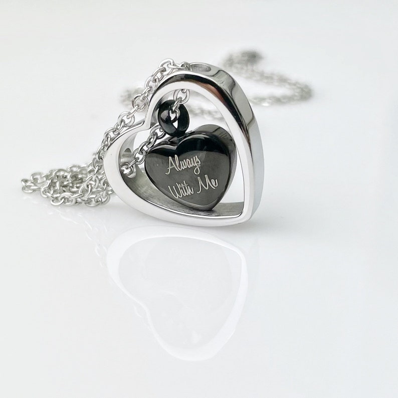 Pode incluir: Um colar com pingente em forma de cora&ccedil;&atilde;o prateado. O pingente tem uma moldura em forma de cora&ccedil;&atilde;o com uma inser&ccedil;&atilde;o em forma de cora&ccedil;&atilde;o preta. A inser&ccedil;&atilde;o &eacute; gravada com as palavras "Always With Me". O colar est&aacute; em uma superf&iacute;cie branca.