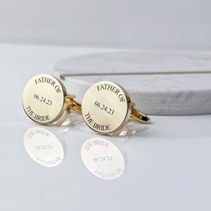 May include: Gold-colored cufflinks with the inscription "FATHER OF THE BRIDE" and the date "06.24.23" engraved on the circular face. The cufflinks are displayed on a reflective surface, showcasing their design.