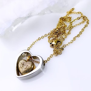 Pode incluir: Um colar dourado com um pingente em forma de cora&ccedil;&atilde;o. O pingente tem um cora&ccedil;&atilde;o externo prateado e um cora&ccedil;&atilde;o dourado menor no interior, com um design de impress&atilde;o digital. A corrente do colar &eacute; dourada e tem um fecho.