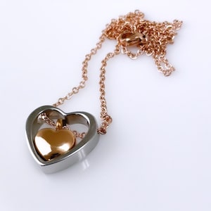 Pode incluir: Um colar com pingente em forma de cora&ccedil;&atilde;o. O cora&ccedil;&atilde;o externo &eacute; prateado, emoldurando um cora&ccedil;&atilde;o menor dourado. A corrente &eacute; de um delicado ouro rosa. O colar &eacute; exibido em um fundo branco.