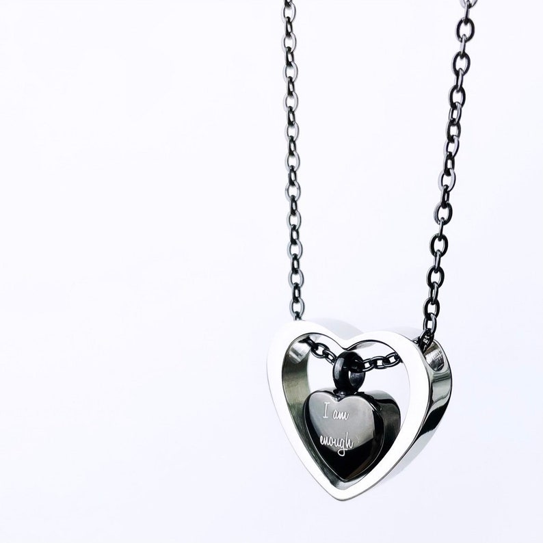 Pode incluir: Um colar com pingente em forma de cora&ccedil;&atilde;o prateado com um pingente de cora&ccedil;&atilde;o preto menor dentro. O pingente de cora&ccedil;&atilde;o preto tem as palavras "I am enough" gravadas. O colar pende de uma corrente escura sobre um fundo branco.
