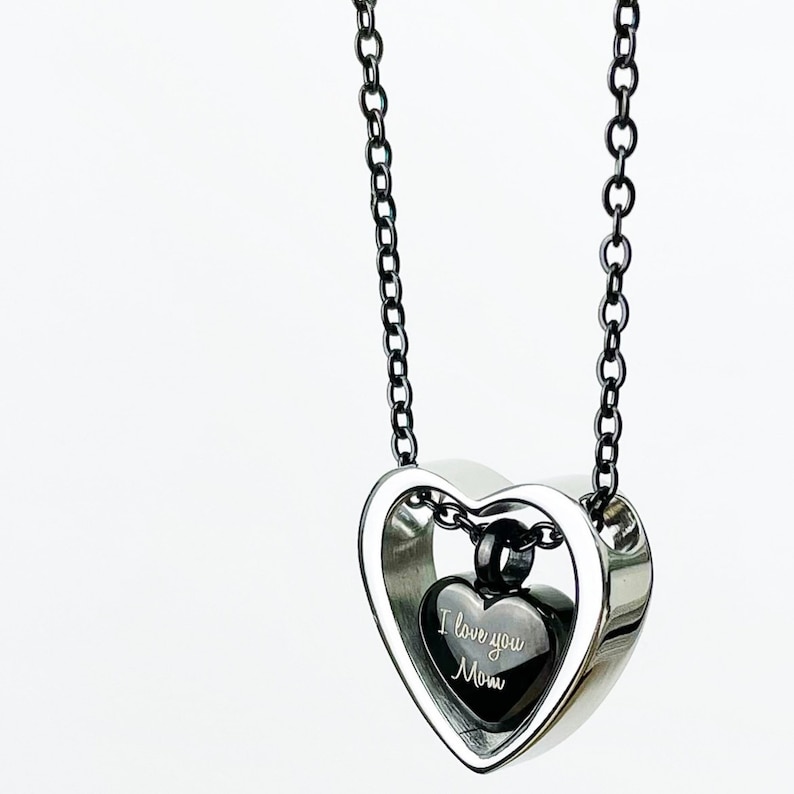 Pode incluir: Um colar com pingente em forma de cora&ccedil;&atilde;o prateado com corrente preta. Dentro do cora&ccedil;&atilde;o maior, h&aacute; um pingente de cora&ccedil;&atilde;o preto menor com a inscri&ccedil;&atilde;o "I love you Mom". O colar est&aacute; em um fundo branco.