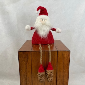 May include: A red and white plush Santa Claus figure with a white beard and a red hat. He is sitting on a wooden surface with his legs dangling down. His legs are made of twine and have pine cone feet.