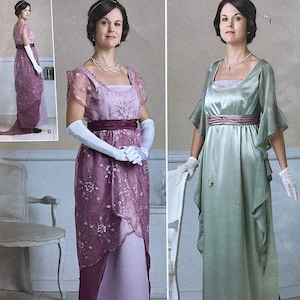 May include: Two women model vintage-style dresses. One dress is a lavender color with a sheer overlay and a burgundy sash. The other dress is a pale green with flowing sleeves and a burgundy sash. Both dresses have a square neckline.
