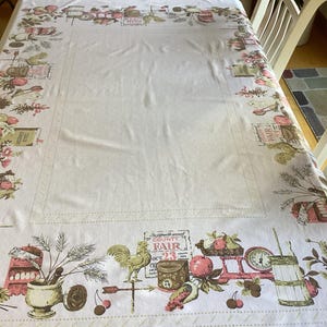 May include: A white rectangular tablecloth with a detailed border featuring a vintage County Fair theme. The border includes illustrations of fruits, vegetables, scales, and a rooster. The tablecloth is set on a wooden table.