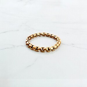 May include: A gold-coloured ring with a chain-link design. The ring is circular and sits on a white surface. The ring's links are rectangular and connected, creating a continuous band. The ring is made of a shiny, metallic material.