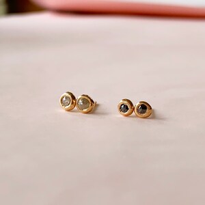 May include: Two pairs of gold stud earrings. One pair features light gray gemstones, and the other pair has black gemstones. The earrings have a simple, elegant design with a bezel setting, perfect for everyday wear.