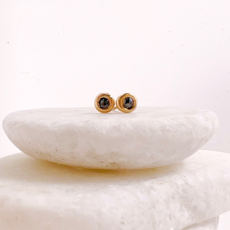 May include: A pair of gold-colored stud earrings, each featuring a dark, round gemstone. The earrings are set against a white background, highlighting their simple yet elegant design. These earrings are a classic jewelry item.