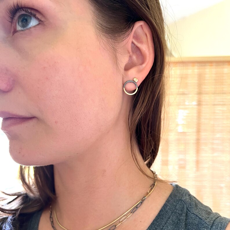 May include: A close-up of a person's ear with a pair of earrings. The earrings feature a gold hoop design with a dark grey top and a small gold stud. A gold and silver chain necklace is also visible.