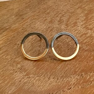 May include: A pair of two-tone stud earrings. Each earring has a dark grey top half and a gold bottom half. The earrings are circular and are presented on a wooden surface. The earrings are a stylish accessory.