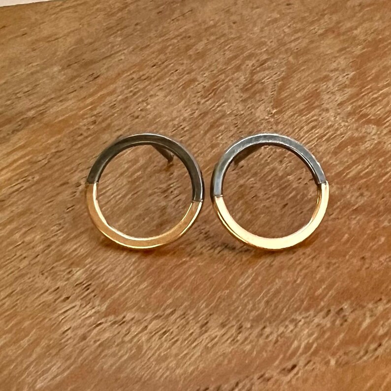 May include: A pair of two-tone stud earrings. Each earring has a dark grey top half and a gold bottom half. The earrings are circular and are presented on a wooden surface. The earrings are a stylish accessory.