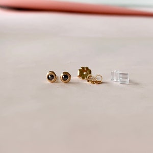 May include: A pair of gold stud earrings with black gemstones. The earrings are round with a bezel setting. Also included are gold earring backs and clear plastic earring stoppers. The earrings are displayed on a light pink surface.