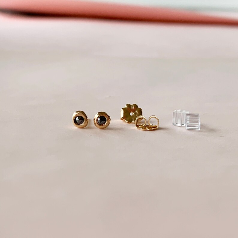 May include: A pair of gold stud earrings with black gemstones. The earrings are round with a bezel setting. Also included are gold earring backs and clear plastic earring stoppers. The earrings are displayed on a light pink surface.
