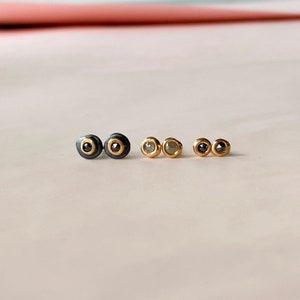 May include: Three pairs of stud earrings in a row. The first pair has a black outer ring with a gold inner ring and a black center stone. The second pair has a gold setting with a light-colored stone. The third pair has a gold setting with a black stone.