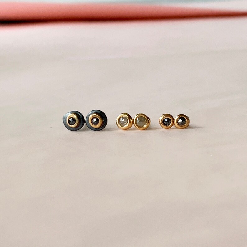 May include: Three pairs of stud earrings in a row. The first pair has a black outer ring with a gold inner ring and a black center stone. The second pair has a gold setting with a light-colored stone. The third pair has a gold setting with a black stone.