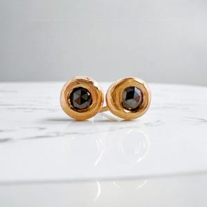 May include: A pair of gold stud earrings, each featuring a dark, round gemstone. The earrings are set in a simple, circular design, with a polished finish. The gemstones have a faceted appearance, catching the light.