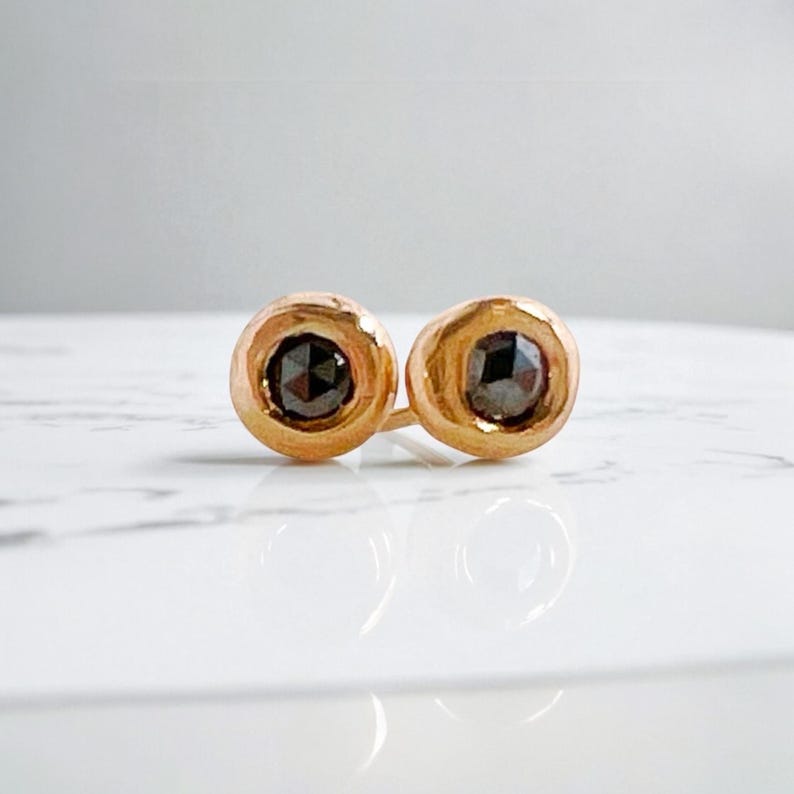 May include: A pair of gold stud earrings, each featuring a dark, round gemstone. The earrings are set in a simple, circular design, with a polished finish. The gemstones have a faceted appearance, catching the light.