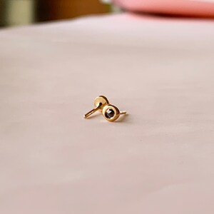 May include: A pair of small, gold-colored stud earrings. Each earring features a small, dark-colored stone set in a bezel. The earrings are displayed on a light pink surface.