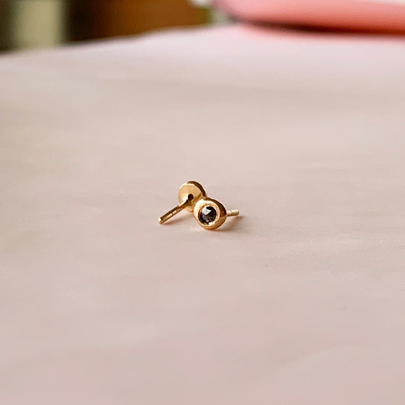May include: A pair of small, gold-colored stud earrings. Each earring features a small, dark-colored stone set in a bezel. The earrings are displayed on a light pink surface.
