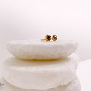 May include: A pair of gold stud earrings with dark gemstones. The earrings are round with a bezel setting, and are displayed on a white, textured surface. The earrings are small and delicate, suitable for everyday wear.