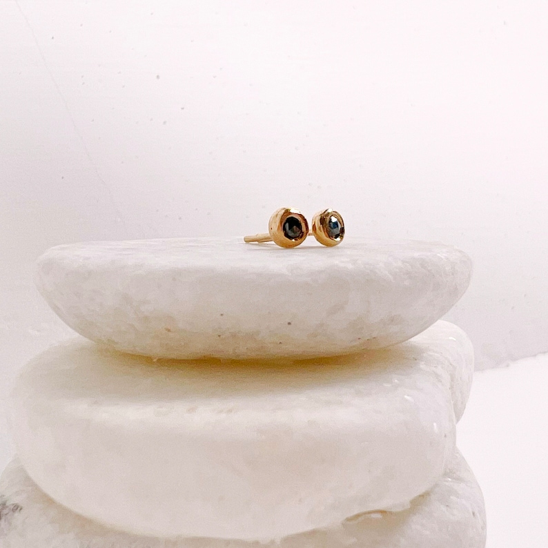 May include: A pair of gold stud earrings with dark gemstones. The earrings are round with a bezel setting, and are displayed on a white, textured surface. The earrings are small and delicate, suitable for everyday wear.