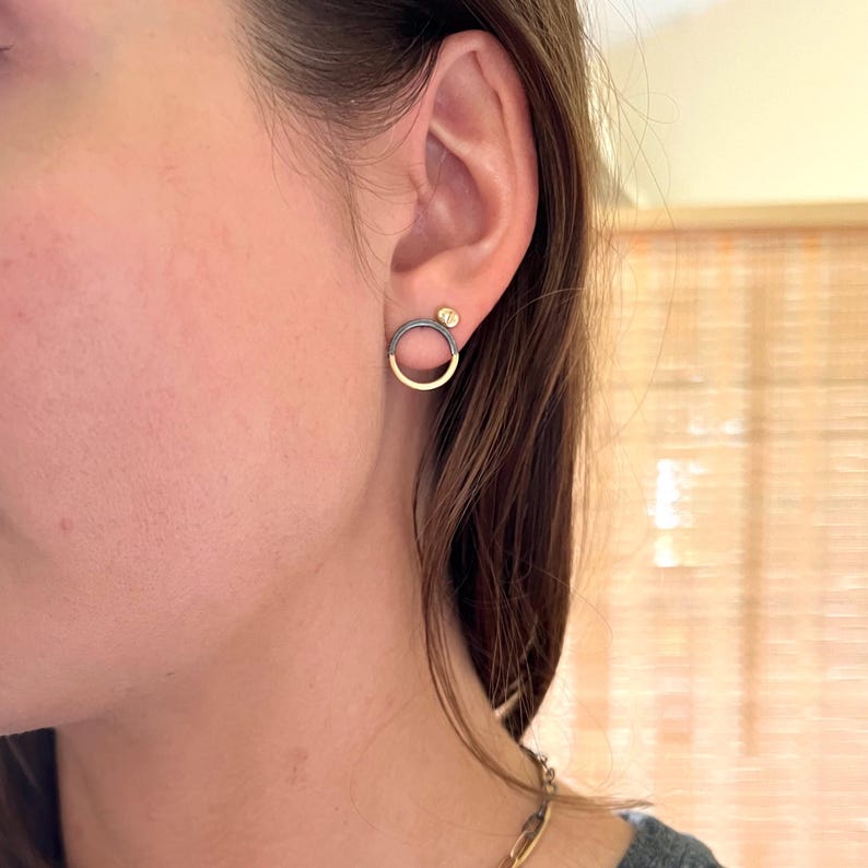 May include: Earrings with a gold and black circular design. One earring has a small, round, gold-coloured accent. The earrings are worn on a person's ear.