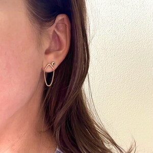 May include: A close-up of a gold earring. The earring features a small, round stud with a black accent, connected to a geometric shape and a delicate chain that dangles below. The earring is worn on a person's ear.