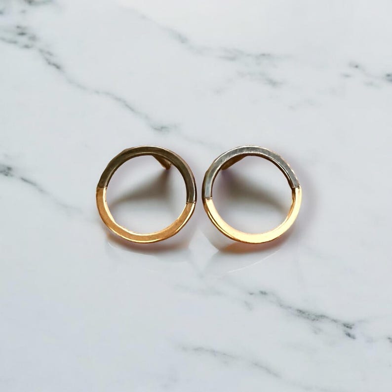 May include: A pair of gold and silver-toned hoop earrings. Each earring is a circle, with the bottom half in gold and the top half in silver. The earrings are displayed on a white marble surface.