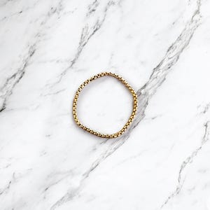 Peut inclure: Un bracelet doré avec un motif de chaîne carrée, posé sur une surface en marbre blanc et gris. Le bracelet est circulaire, avec de petits maillons rectangulaires. L'esthétique générale est minimaliste et élégante.