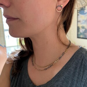 May include: A person wearing two necklaces and earrings. The earrings are a gold circle with a dark metal accent and a small gold ball. The necklaces are gold and dark metal chain styles.