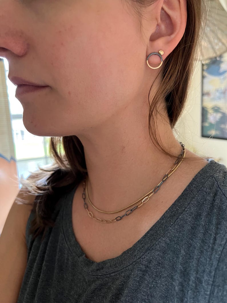 May include: A person wearing two necklaces and earrings. The earrings are a gold circle with a dark metal accent and a small gold ball. The necklaces are gold and dark metal chain styles.
