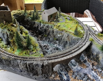 Puede incluir: Un detallado paisaje de tren en miniatura con una vía sinuosa, un puente de piedra y edificios en miniatura. La escena incluye un río, acantilados rocosos y pequeños árboles de hoja perenne, creando un entorno en miniatura realista.