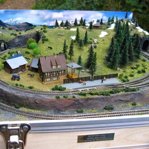 May include: A detailed model railway scene featuring a mountain landscape with a winding track, tunnels, miniature buildings, trees, and figures. The scene includes a station, houses, and a farm, all set against a backdrop of mountains and sky.